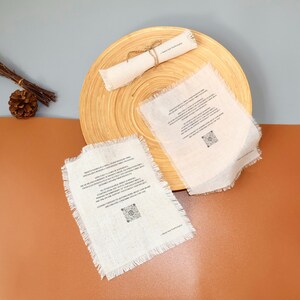 May include: A natural bamboo plate holds two white linen napkins with frayed edges and printed text. A rolled linen scroll tied with twine rests on the plate. The background is a mix of brown and gray.