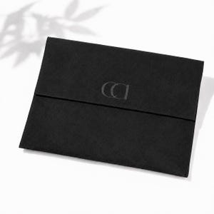 Könnte beinhalten: Eine rechteckige, schwarze Tasche im Umschlag-Stil mit einer glatten, matten Textur. Die vordere Klappe zeigt ein zentriertes, geprägtes Logo, das aus den ineinander verschlungenen Buchstaben CA in einer serifenlosen Schrift besteht.