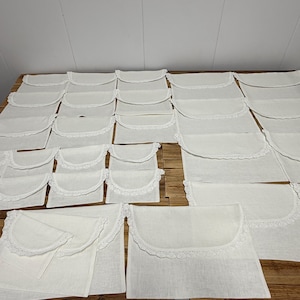 May include: A collection of off-white fabric pouches arranged in rows. Each rectangular pouch features a rounded flap closure adorned with a delicate white lace trim along the curved edge. The pouches are made of a plain, light-colored textile.