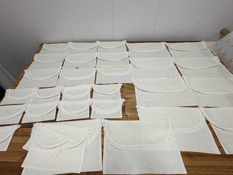 May include: A collection of off-white fabric pouches arranged in rows. Each rectangular pouch features a rounded flap closure adorned with a delicate white lace trim along the curved edge. The pouches are made of a plain, light-colored textile.