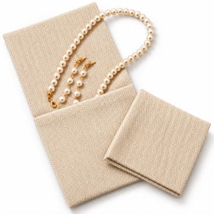 Puede incluir: Una bolsa de tela beige para joyas con patrón de espiga, un cierre dorado y un collar de perlas color crema en su interior. Una pieza de tela cuadrada a juego con la misma textura de espiga se encuentra colocada al lado.