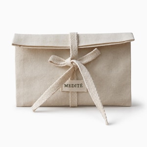Peut inclure: Une pochette rectangulaire en toile beige, nouée avec un ruban et un nœud assortis. La pochette a un dessus replié et une petite étiquette avec le mot "MEDITE" imprimé. L'esthétique générale est minimaliste et naturelle.