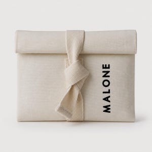 Puede incluir: Una bolsa rectangular de lona blanquecina con la parte superior enrollada y un cierre de correa anudada. La palabra "MALONE" está impresa verticalmente en negro en la parte delantera. La bolsa está hecha de tela texturizada.