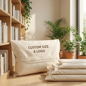 May include: Cream-colored pillow with the text "CUSTOM SIZE & LOGO" in brown. The pillow has a folded flap and a strap. Several smaller pillows are stacked in front of the larger pillow. The pillows are in a room with plants and a bookshelf.