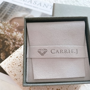 May include: A gray velvet jewelry box with a gray suede interior. The box has a diamond logo and the text "CARRIE.J" embossed on the front.