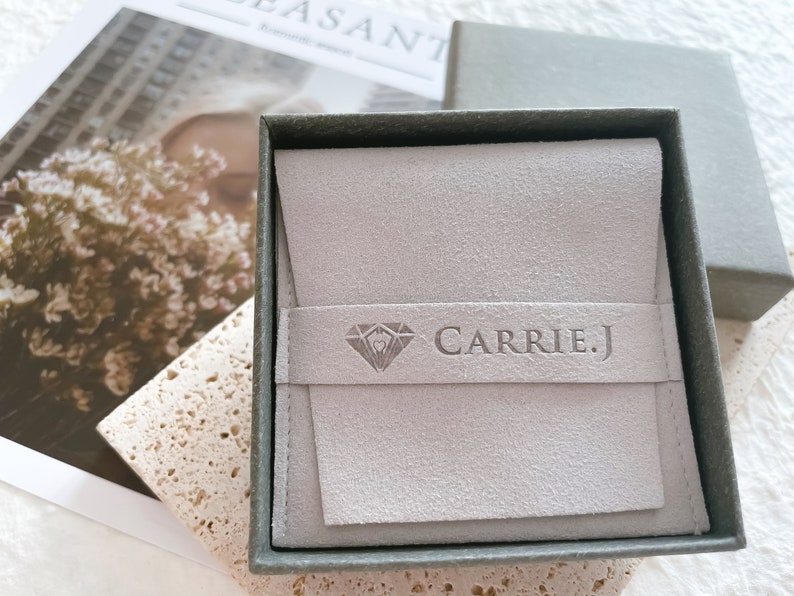 May include: A gray velvet jewelry box with a gray suede interior. The box has a diamond logo and the text "CARRIE.J" embossed on the front.