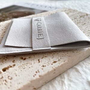 May include: A white, folded, rectangular pouch with the text "CARRIE J." embossed on the front.
