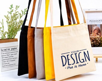 May include: A collection of canvas tote bags in various colors, including black, brown, gray, and yellow. The bags have long straps and the front of one bag displays the text "YOUR DESIGN - Put It Here" in navy blue. A paper bag with text is in the background.