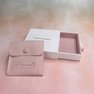 Puede incluir: Una caja blanca con un caj&oacute;n que se desliza hacia afuera. La caja est&aacute; etiquetada con "TreePackaging". El caj&oacute;n est&aacute; forrado con terciopelo rosa y contiene una bolsa de terciopelo rosa con el texto "Howard Health & Healing" en blanco.