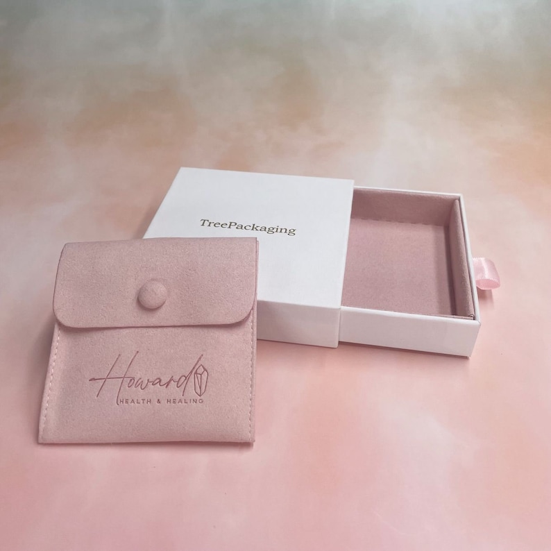 Puede incluir: Una caja blanca con un caj&oacute;n que se desliza hacia afuera. La caja est&aacute; etiquetada con "TreePackaging". El caj&oacute;n est&aacute; forrado con terciopelo rosa y contiene una bolsa de terciopelo rosa con el texto "Howard Health & Healing" en blanco.