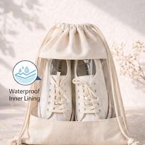 May include: A beige drawstring bag made of canvas fabric featuring a clear plastic window on the front. Inside the bag, a pair of white lace-up trainers are visible. The bag includes a graphic icon and the text Waterproof Inner Lining.