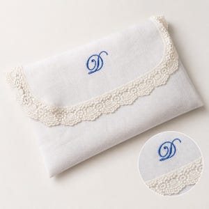 May include: A white rectangular fabric envelope pouch featuring a scalloped cream-colored lace trim along the flap edge. The center of the flap is embroidered with a blue cursive letter D. A circular inset shows a detailed view of the embroidery.