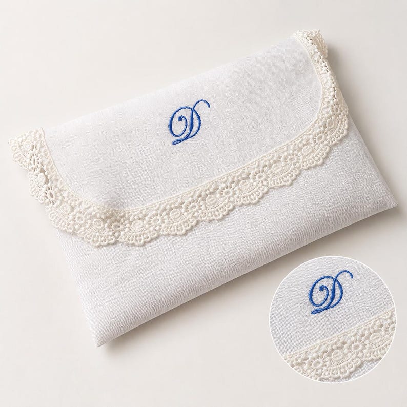 May include: A white rectangular fabric envelope pouch featuring a scalloped cream-colored lace trim along the flap edge. The center of the flap is embroidered with a blue cursive letter D. A circular inset shows a detailed view of the embroidery.
