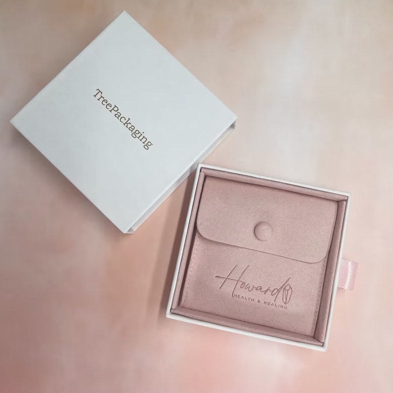 Puede incluir: Una caja blanca con el texto "TreePackaging" impreso en la parte superior. La caja est&aacute; abierta y revela una bolsa de terciopelo rosa con el texto "Howard Health & Healing" impreso en ella.