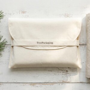 May include: A cream-colored, rectangular package with a curved flap and a matching strap, bearing the text "TreePackaging". A folded stack of fabric is to the right. A pine branch with cones rests on a white wooden surface.