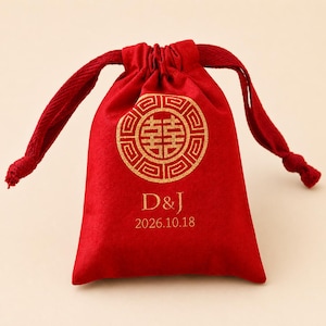 以下が含まれることがあります： 金色の円形デザインとイニシャル「D&J」、日付「2026.10.18」が金色でプリントされた赤い巾着袋。袋は柔らかい素材でできており、お揃いの赤い巾着開閉部が付いています。