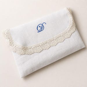 May include: A white rectangular fabric pouch featuring a curved flap edge trimmed with intricate cream-colored lace. The center of the flap displays a single embroidered letter D in a vibrant blue thread, centered on the plain white fabric surface.