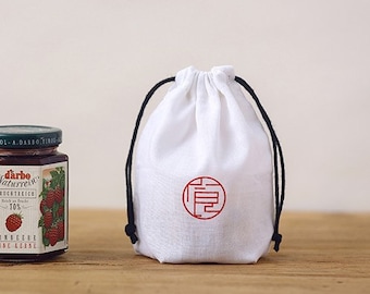 May include: A white drawstring bag with a black drawstring and a red Chinese character on the front. The bag is sitting on a wooden surface next to a jar of raspberry jam.