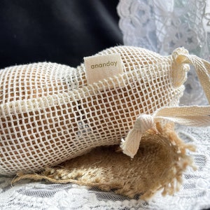 May include: A beige mesh bag with a drawstring closure and a label that says "ananday". The bag is tied with a white ribbon and is sitting on a white lace surface.