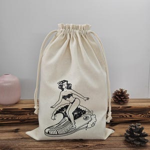 May include: A cream-coloured drawstring bag with a black and white illustration of a woman surfing on a shoe-shaped wave. The bag has a gathered top and a drawstring closure.