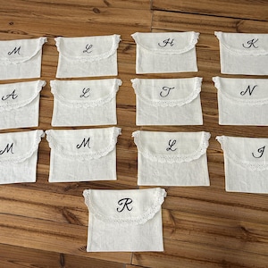 May include: A collection of thirteen off-white rectangular fabric pouches with envelope-style flaps. Each flap features a scalloped lace trim and a single embroidered black letter in a cursive script, including the letters M, L, H, K, A, T, N, J, and R.