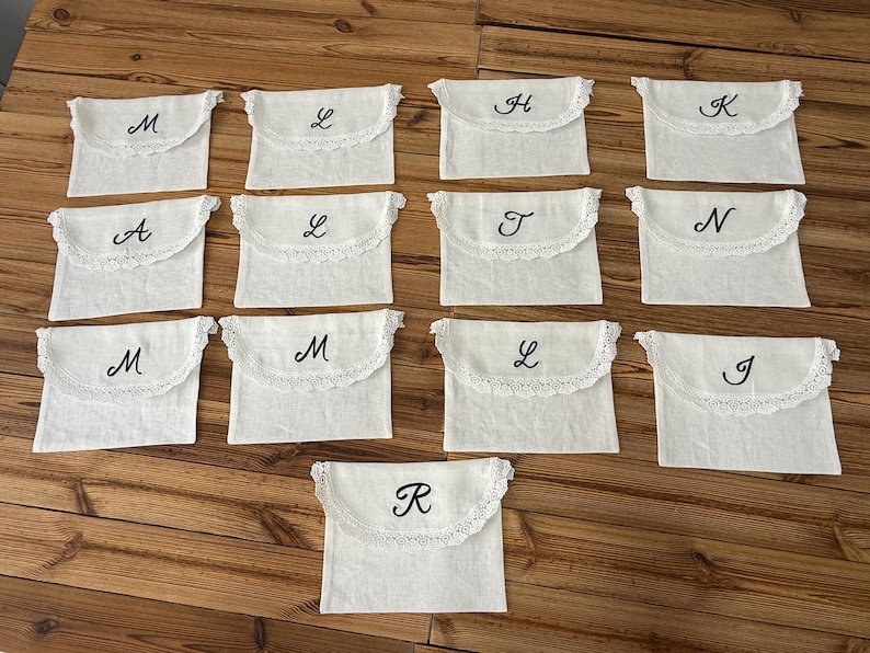 May include: A collection of thirteen off-white rectangular fabric pouches with envelope-style flaps. Each flap features a scalloped lace trim and a single embroidered black letter in a cursive script, including the letters M, L, H, K, A, T, N, J, and R.