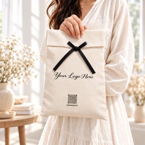 Puede incluir: Una bolsa rectangular de tela color hueso con una cinta negra atada en un lazo en la parte superior. El frente muestra el texto negro Your Logo Here, un código QR cuadrado y el nombre de usuario @TREEPACKAGING.ETSY impresos en tinta negra.