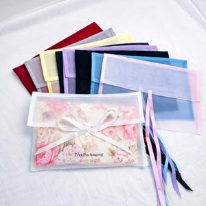 Può includere: Una collezione di sacchetti trasparenti colorati con nastri coordinati. Un sacchetto contiene un articolo con stampa floreale ed è legato con un nastro bianco. I sacchetti sono in vari colori, tra cui rosso, giallo, blu e nero. Il testo "TreePackaging" è visibile.