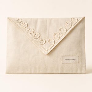 Op de afbeelding: Een crèmekleurig stoffen enveloptasje met een driehoekige flap versierd met decoratief koordborduurwerk in krulpatronen langs de randen. Een klein rechthoekig stoffen label in de rechterbenedenhoek toont de tekst TreePackaging.