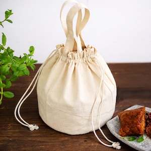 Peut inclure: Un sac en toile de couleur crème avec un cordon de serrage, un haut froncé et deux poignées. Le sac est rond et posé sur une surface en bois. Une assiette de nourriture est à droite, et des plantes vertes à feuilles sont à gauche.
