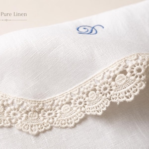 May include: A white linen textile featuring the text 100% Pure Linen, adorned with a beige crocheted lace trim along the curved edge and a small blue embroidered letter D monogram centered on the fabric surface.