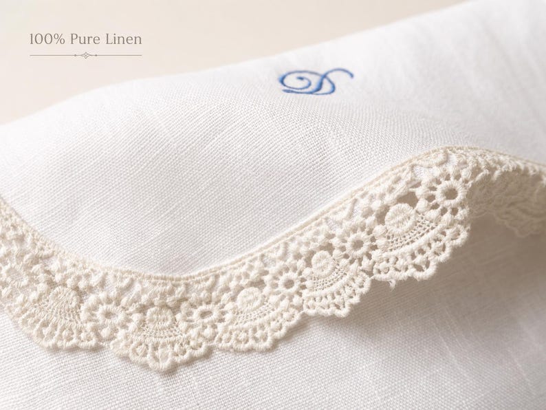 May include: A white linen textile featuring the text 100% Pure Linen, adorned with a beige crocheted lace trim along the curved edge and a small blue embroidered letter D monogram centered on the fabric surface.