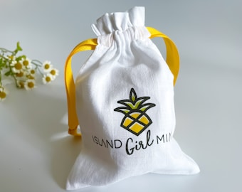 May include: White drawstring bag with yellow ribbon ties. The bag has a pineapple graphic and the text "Island Girl Mini" printed on the front.