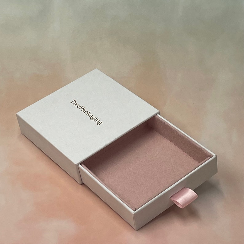 Puede incluir: Caja de caj&oacute;n blanca con interior rosa y un tirador de cinta rosa. La caja tiene el texto "TreePackaging" impreso en la parte superior.