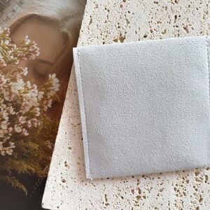 May include: A light gray suede pouch with a smooth texture. The pouch is lying on a light beige textured surface.