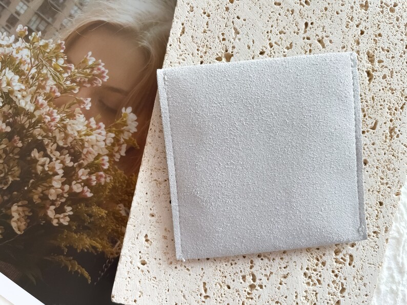 May include: A light gray suede pouch with a smooth texture. The pouch is lying on a light beige textured surface.