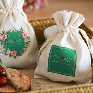 Peut inclure: Deux petits sacs en tissu blanc à cordon avec des motifs verts et floraux. Un sac dit "Just Married" et l'autre dit "Just Married". Les sacs sont dans un panier en osier.