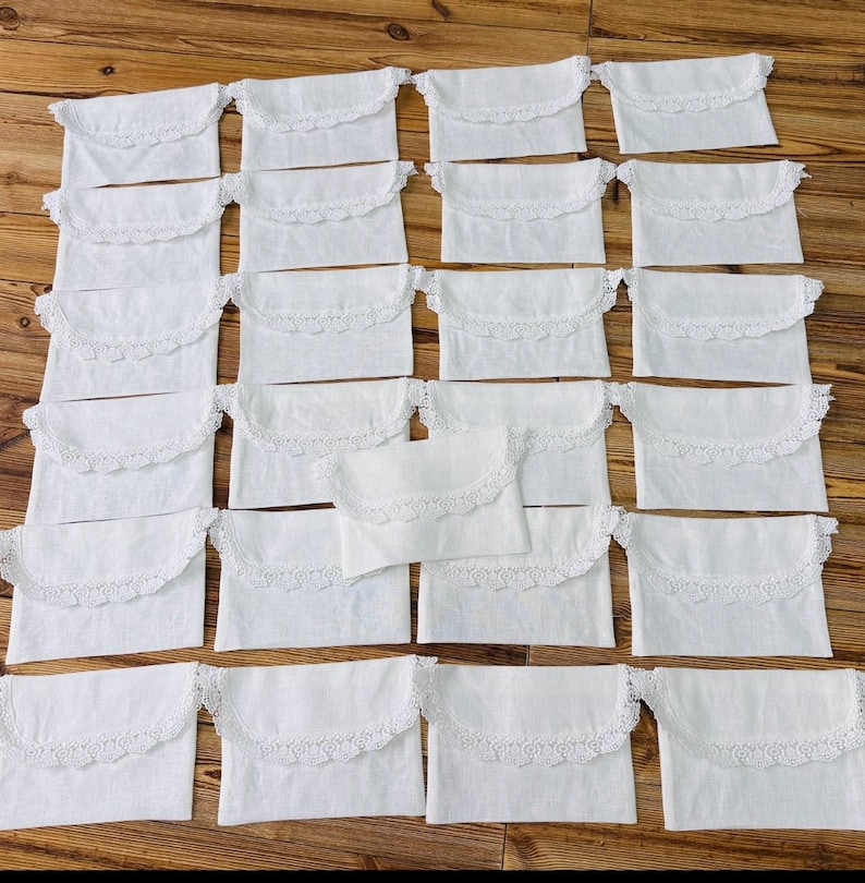 May include: A collection of twenty-one rectangular white fabric pouches, each featuring a scalloped flap edge trimmed with delicate white lace. The pouches are arranged in a grid pattern, showcasing a uniform design with a soft, woven texture.