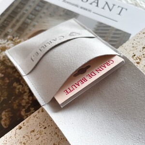 May include: A white suede pouch with a flap that has the word "CARRIE" embossed on it. The pouch is open and a small card with the text "GRAIN DE BEAUTE" is visible inside.