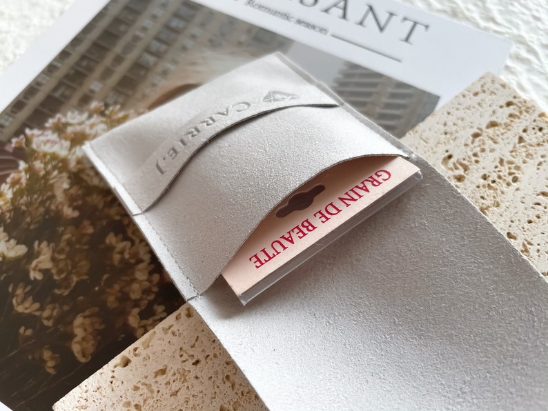 May include: A white suede pouch with a flap that has the word "CARRIE" embossed on it. The pouch is open and a small card with the text "GRAIN DE BEAUTE" is visible inside.