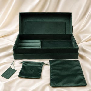 Puede incluir: Un joyero de terciopelo verde oscuro con la tapa abierta, que cuenta con un inserto para anillos y un compartimento de almacenamiento plano. Incluye una pequeña bolsa con cordón, un paño rectangular y una etiqueta, todo en terciopelo verde.