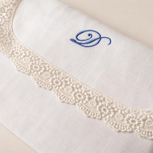 May include: A white fabric envelope-style pouch featuring a scalloped cream-colored lace trim along the flap edge. The front of the pouch is decorated with a single embroidered letter D in a cursive blue thread, centered above the lace border.