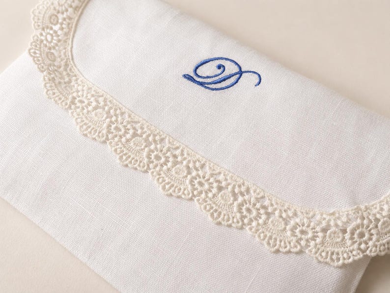 May include: A white fabric envelope-style pouch featuring a scalloped cream-colored lace trim along the flap edge. The front of the pouch is decorated with a single embroidered letter D in a cursive blue thread, centered above the lace border.