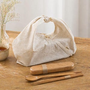 May include: A beige fabric lunch bag with a knotted handle, a wooden utensil case, a wooden spoon, and chopsticks. The bag and utensil case are secured with a beige band. The items are on a wooden surface.