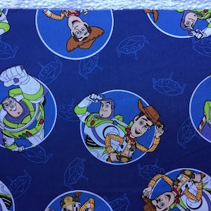 May include: Blue fabric with a repeating pattern of Woody and Buzz Lightyear from Toy Story. The characters are in circles on a blue background with a pattern of flying saucers.