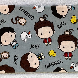 Może przedstawiać: Szary materiał z powtarzającym się wzorem postaci z kreskówek z serialu Friends. Postacie to Joey, Chandler i Ross. Materiał zawiera również kreskówkowe zwierzęta, w tym małpy, kaczki i żółtą gumową kaczkę. Materiał ma nadrukowane słowa "Joey", "Chandler" i "Ross".