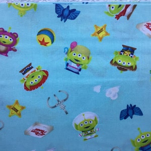 Puede incluir: Una tela azul con un patrón repetido de alienígenas verdes de tres ojos en varias poses, incluyendo el uso de un sombrero de vaquero, un sombrero y un traje espacial. La tela también presenta una estrella amarilla, una pelota azul y amarilla, y una nave espacial azul y blanca.