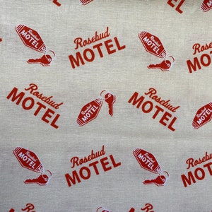 Könnte beinhalten: Ein beigefarbener Stoff mit einem sich wiederholenden Muster aus rotem "Rosebud Motel"-Text und einem Schlüssel mit einem Motel-Schlüsselanhänger.