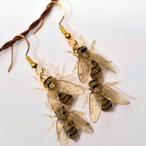 May include: A pair of dangle earrings featuring three translucent, glitter-infused bee charms suspended from gold-toned metal hooks. Each bee charm has a clear body with visible black stripes and delicate wings, creating a layered, insect-themed design.