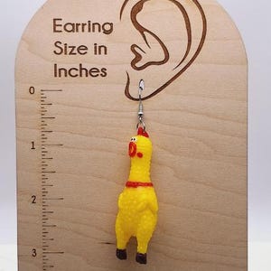 May include: A yellow rubber chicken earring with a red comb and feet, hanging from a silver-toned metal hook. The earring is displayed on a light-colored wooden card featuring an ear outline and a ruler marked in inches, showing the chicken is approximately 2.5 inches long.